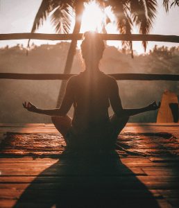 Person meditating outdoors at sunrise with palm trees in the background.